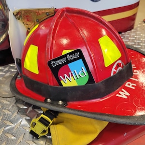 May include: A red fire helmet with a yellow reflective patch and a "Draw four Wild" card sticker attached to the front.