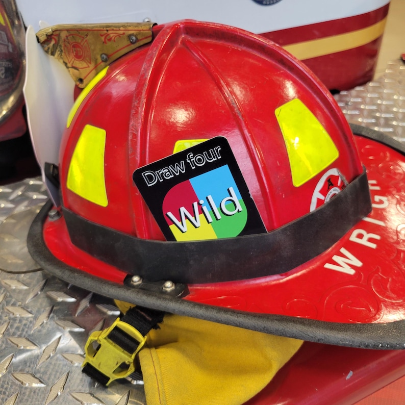 May include: A red fire helmet with a yellow reflective patch and a "Draw four Wild" card sticker attached to the front.