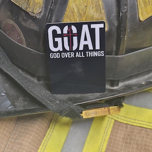 May include: A rectangular black sticker featuring the white text GOAT with a red cross integrated into the letter O, and the white text GOD OVER ALL THINGS underneath, positioned on a black and yellow firefighter helmet with reflective trim.