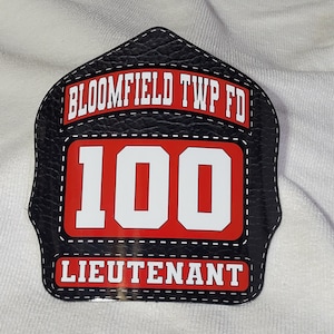 May include: Black leather firefighter helmet patch with white stitching. The patch has red lettering that says "Bloomfield Twp FD" and "100" and "Lieutenant".
