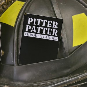 May include: A black and white card with the text "Pitter Patter Throw a Ladder" on a black background. The card is attached to a black helmet with yellow accents.