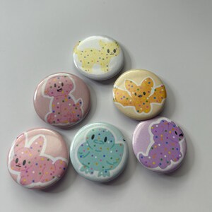 May include: Six colorful round buttons with different animal designs. Each button features a different animal, including a dog, a cat, a bat, a chick, a bunny, and a dinosaur, all with sprinkles on them.