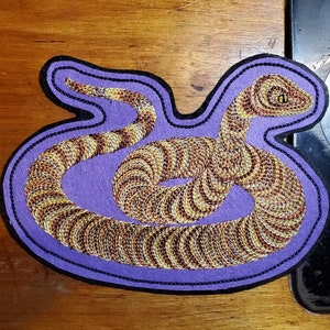 May include: Embroidered snake patch with a detailed design in brown, tan, and yellow hues, set against a purple background with a black border. The patch is on a wooden surface, near a sewing machine.