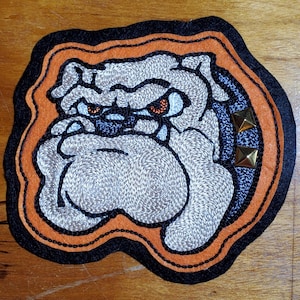 May include: An embroidered patch featuring a grey bulldog head with orange eyes, framed by a thick orange and black border. The design includes a dark blue collar section adorned with two small, square, metallic gold-colored studs on the right side.