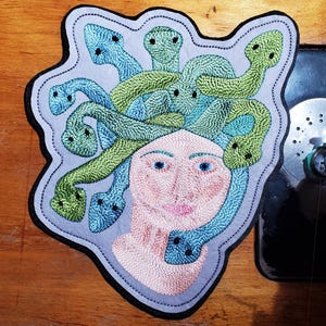 May include: Embroidered patch featuring Medusa with blue eyes, pink lips, and a head of green and blue snakes. The patch has a light blue background and a black border. The patch is on a wooden surface.