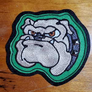 May include: An embroidered patch featuring a grey bulldog head with orange eyes and a dark blue collar with silver studs. The design is outlined in a bright green border, which is then framed by a thick black border, creating a layered effect.