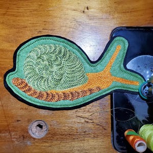 May include: Embroidered snail applique in shades of green, orange, and brown, with a black border. The snail is on a wooden surface next to a vintage sewing machine and spools of thread. Craft project.