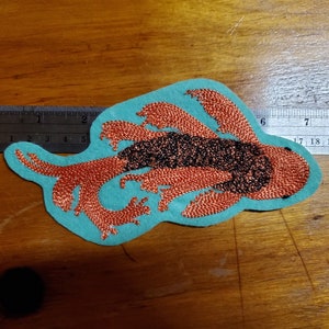 May include: An embroidered patch featuring a stylized lobster design. The lobster is rendered in copper-colored thread with a black central body, set against a light blue felt backing. The patch measures approximately 5 inches in length.