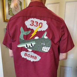 May include: A maroon short-sleeved shirt with an embroidered design on the back. The design features a green airplane with a lightning bolt and the number 330 in a cloud. The words "BOMB" are also embroidered in a cloud.