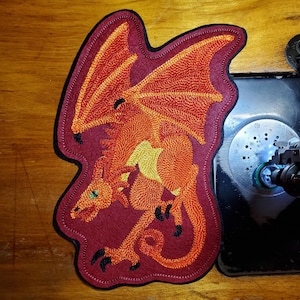 May include: A red, orange, and yellow embroidered dragon patch with black claws. The patch is on a brown wooden surface.