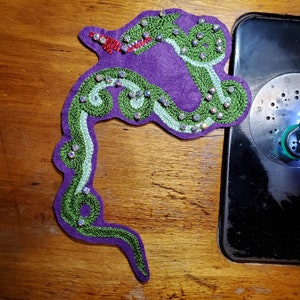 May include: A purple felt applique in the shape of a snake, with green embroidered details and iridescent rhinestones. The snake has a red tongue and is lying on a wooden surface. A sewing machine is visible in the background.