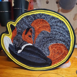 May include: A black, orange, and white embroidered patch of a curled up fox with a yellow border.