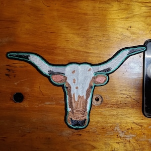 May include: Embroidered patch of a white and brown longhorn steer head with turquoise horns. The patch is on a wooden surface.