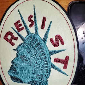 May include: A white oval patch with a red embroidered outline. The center of the patch features a blue and black embroidered image of the Statue of Liberty's head. The words "RESIST" are embroidered in red around the image.
