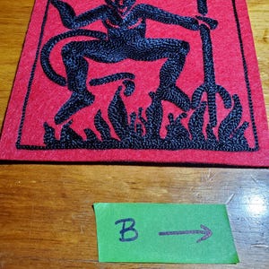 Op de afbeelding: Een vierkante rode vilten patch met zwart borduurwerk van een gehoornde figuur die een drietand boven vlammen vasthoudt. Onder de patch ligt een klein groen rechthoekig papieren label met de handgeschreven letter B en een pijl naar rechts.