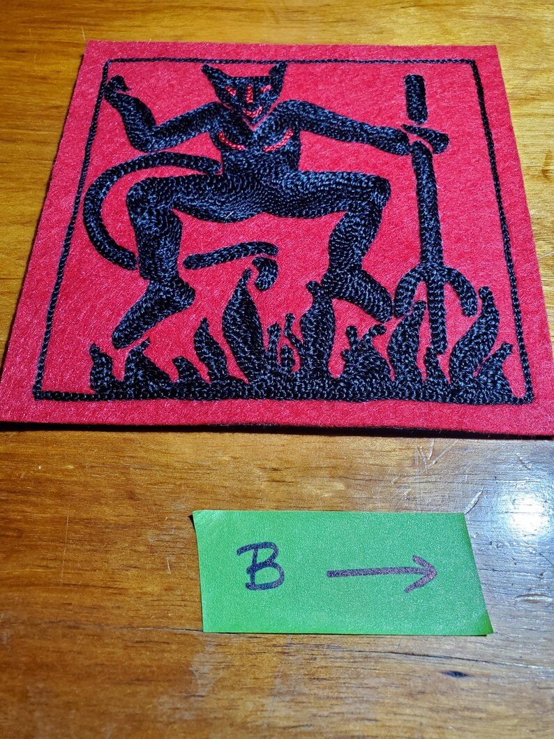 Op de afbeelding: Een vierkante rode vilten patch met zwart borduurwerk van een gehoornde figuur die een drietand boven vlammen vasthoudt. Onder de patch ligt een klein groen rechthoekig papieren label met de handgeschreven letter B en een pijl naar rechts.