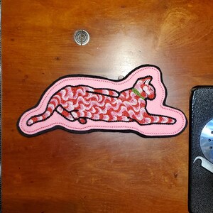 May include: An embroidered patch shaped like a reclining cat with a pink background and a red and white wavy striped pattern on its body. The cat has a small green collar and is outlined with a thick black border, featuring detailed stitching.