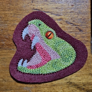 May include: An embroidered patch on a dark maroon felt backing featuring a green snake head with its mouth open. The snake has a bright orange eye with a black pupil, white fangs, and a pink interior mouth, all rendered in detailed thread work.