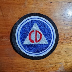 May include: A circular embroidered patch featuring a black outer border, a white inner ring, and a blue circular field. Centered within the blue field is a light gray triangle containing the red embroidered letters CD in a bold, sans-serif font.