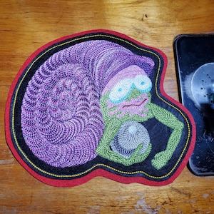 May include: Embroidered patch featuring a whimsical creature with a purple spiral head, green body, and blue eyes. The patch has a black border with yellow and red accents. The design is on a wooden surface.