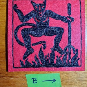 Op de afbeelding: Een vierkante rode viltpatch met zwarte machinale borduursels van een gehoornde figuur met een staart, die een drietand boven vlammen vasthoudt. De figuur heeft kleine rode gestikte accenten op het gezicht en de borst. Een groen papieren label met tekst B en pijl.