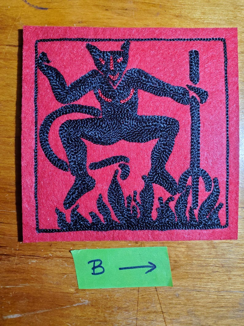 Op de afbeelding: Een vierkante rode viltpatch met zwarte machinale borduursels van een gehoornde figuur met een staart, die een drietand boven vlammen vasthoudt. De figuur heeft kleine rode gestikte accenten op het gezicht en de borst. Een groen papieren label met tekst B en pijl.