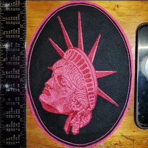 May include: A pink embroidered patch featuring the Statue of Liberty's head and crown, set against a black oval background with a pink border. The patch is likely for clothing or accessories.