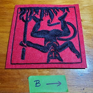 Op de afbeelding: Een vierkante rode stoffen patch met zwarte borduursels van een omgekeerde figuur die een drietand vasthoudt. De figuur heeft een staart en puntige oren, met kleine rode stiksels. Onder de patch bevindt zich een groen label met de tekst B &rarr;.