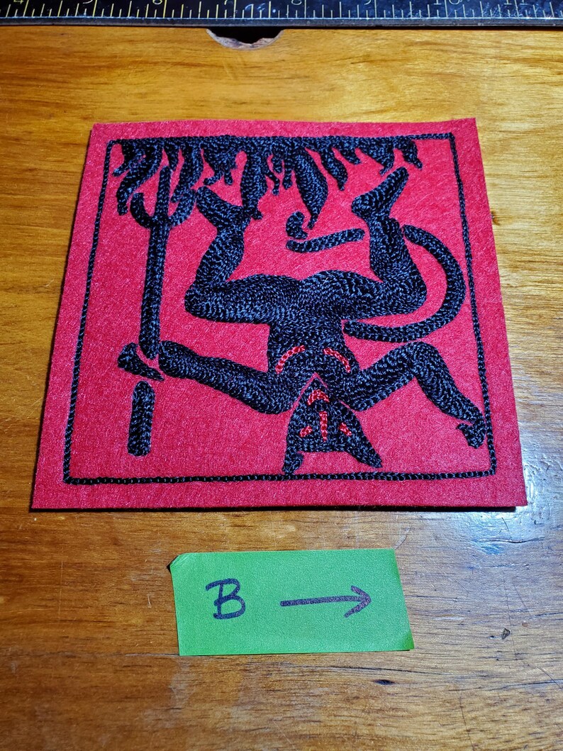 Op de afbeelding: Een vierkante rode stoffen patch met zwarte borduursels van een omgekeerde figuur die een drietand vasthoudt. De figuur heeft een staart en puntige oren, met kleine rode stiksels. Onder de patch bevindt zich een groen label met de tekst B &rarr;.