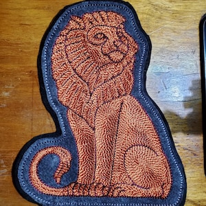 May include: Embroidered patch featuring a seated lion in shades of orange and brown. The lion is detailed with textured stitching, set against a dark gray felt background with a stitched border. The patch is a unique decorative item.