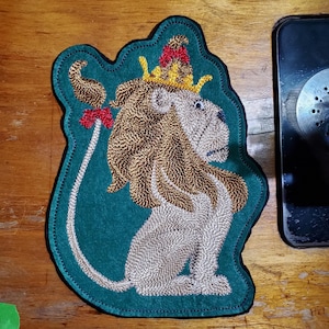 May include: Embroidered patch featuring a lion wearing a crown. The lion is beige with a brown mane and tail, set against a teal background. The patch has a decorative border and a red bow. This patch is suitable for jackets, bags, or other fabric items.