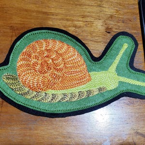 May include: A snail-shaped patch with an orange and yellow shell, green body, and black border. The snail's body is a light green color, and the shell is a detailed, textured design. The patch is likely for clothing or accessories.