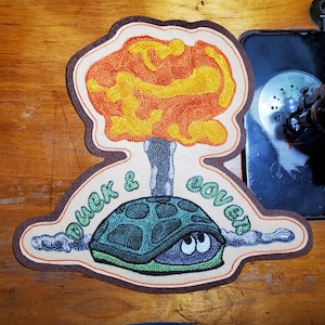 May include: An embroidered patch featuring a teal turtle with visible eyes hiding under its shell. Above the turtle is a large, textured orange and yellow explosion cloud. The text "DUCK & COVER" is stitched in green letters around the turtle.