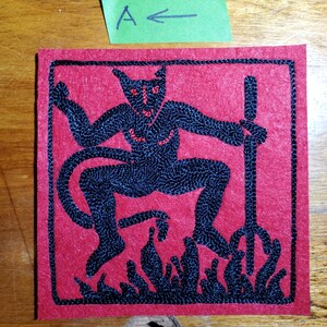 Op de afbeelding: Een vierkante rode viltpatch met zwarte machinale borduurwerk van een gehoornd figuur met een drietand, staand boven vlammen. Het figuur heeft rode ogen en borstdetails. Een groen papieren label erboven toont de tekst A met een pijl.