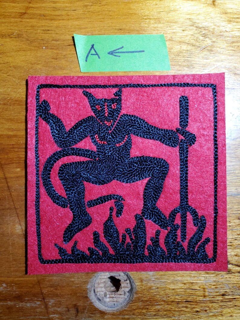 Op de afbeelding: Een vierkante rode viltpatch met zwarte machinale borduurwerk van een gehoornd figuur met een drietand, staand boven vlammen. Het figuur heeft rode ogen en borstdetails. Een groen papieren label erboven toont de tekst A met een pijl.