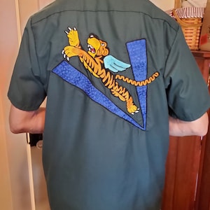 May include: A dark green short-sleeved button-down shirt with an embroidered design on the back. The design features a yellow and orange tiger with blue wings, flying over a blue and black geometric shape.