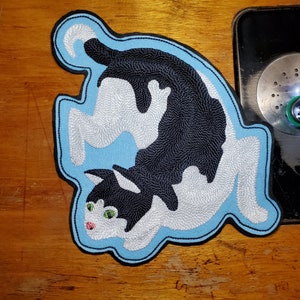 May include: A black and white embroidered cat patch on a light blue background. The cat is curled up in a playful pose with its tail curled around its body.