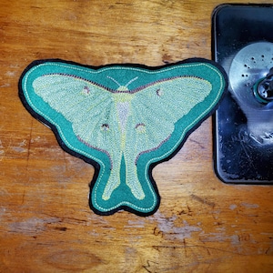 May include: A large, embroidered moth patch with a teal outline and a black border. The moth's wings are light green with detailed stitching in yellow and maroon. The patch is on a wooden surface near a sewing machine.