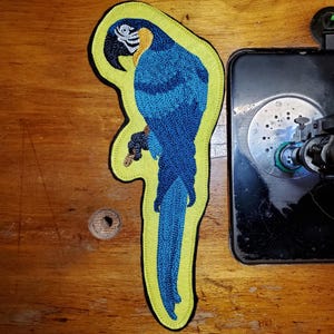 May include: A blue and yellow embroidered patch of a parrot perched on a branch. The patch is outlined in yellow stitching.