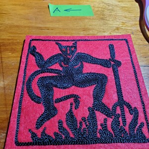 Op de afbeelding: Een vierkante rode stoffen patch met zwart borduurwerk van een gehoornde figuur die een drietand boven vlammen vasthoudt. De patch ligt onder een metalen liniaal met cm-aanduiding en een groen label met de letter A.