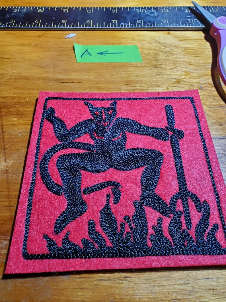 Op de afbeelding: Een vierkante rode stoffen patch met zwart borduurwerk van een gehoornde figuur die een drietand boven vlammen vasthoudt. De patch ligt onder een metalen liniaal met cm-aanduiding en een groen label met de letter A.