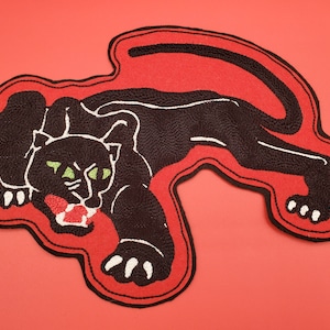 May include: Black panther patch with green eyes and a red mouth, set against a red background. The panther is embroidered with black thread and outlined in a red border. The patch is a unique accessory.