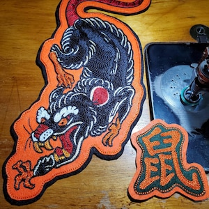 May include: An embroidered patch featuring a black rat with orange accents and a long tail, alongside a smaller patch with the orange Chinese character for rat on a green background, both outlined in orange thread on a black border.