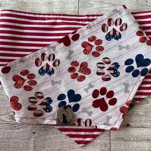 Op de afbeelding: Een rood, wit en blauwe hondenbandana met een pootprintpatroon en een botontwerp. De bandana is omkeerbaar met een rood-wit gestreept patroon aan de andere kant.