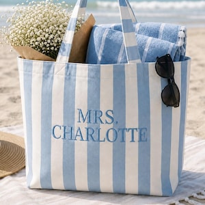 Pode incluir: Uma bolsa tote com listras verticais em azul claro e branco, apresentando o texto bordado MRS. CHARLOTTE em azul. A bolsa contém uma toalha listrada em azul e branco e um buquê de flores brancas, com óculos de sol pretos anexados.