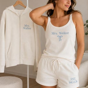 Puede incluir: Una sudadera con capucha y cremallera color crema junto a un conjunto de camiseta sin mangas y pantalones cortos. La sudadera tiene bordado "Mrs. Walker" en azul. La camiseta muestra "Mrs. Walker" y un lazo azul, el pantalón "EST. 04.12.26".