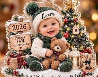 May include: A clear acrylic Christmas ornament featuring a baby in a green knit hat and scarf holding a teddy bear. The design includes a sign reading 2026 Baby's First Christmas, a decorated tree, gifts, and the text A little boy, a big blessing.