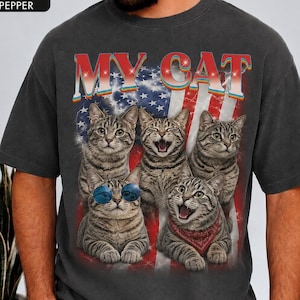 May include: A charcoal gray t-shirt featuring a graphic print of five tabby cats against a background of the American flag. The text MY CAT is printed in a rainbow gradient font above the cats. One cat wears blue sunglasses and another a red bandana.
