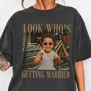Könnte beinhalten: Ein anthrazitfarbenes Kurzarm-T-Shirt mit einer zentralen rechteckigen Grafik eines Kindes mit Sonnenbrille und gestreiftem Tanktop, das auf einer Bank sitzt. Der Text lautet LOOK WHO'S GETTING MARRIED mit dem Namen Emma in Schreibschrift.
