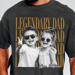 Pode incluir: Camiseta cinza escuro com uma foto em preto e branco de duas crianças usando óculos de sol. As palavras "LEGENDARY DAD" são impressas em dourado, repetidas verticalmente. Os nomes "Liam & Olivia" são impressos abaixo.